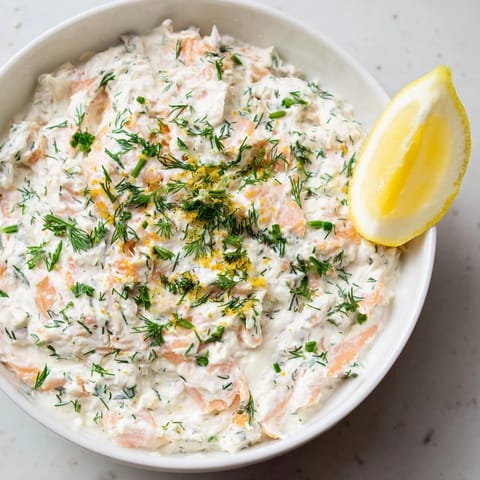 This elegant smoked salmon dip sits in a white bowl, surrounded by toasted bagel chips and cucumber slices.