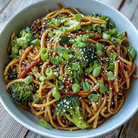 Bright orange carrots and green broccoli florets are tossed with tender egg noodles in a glossy teriyaki sauce in a ceramic bowl. Toasted sesame seeds and sliced green onions garnish the Asian Teriyaki Noodle Bowl.