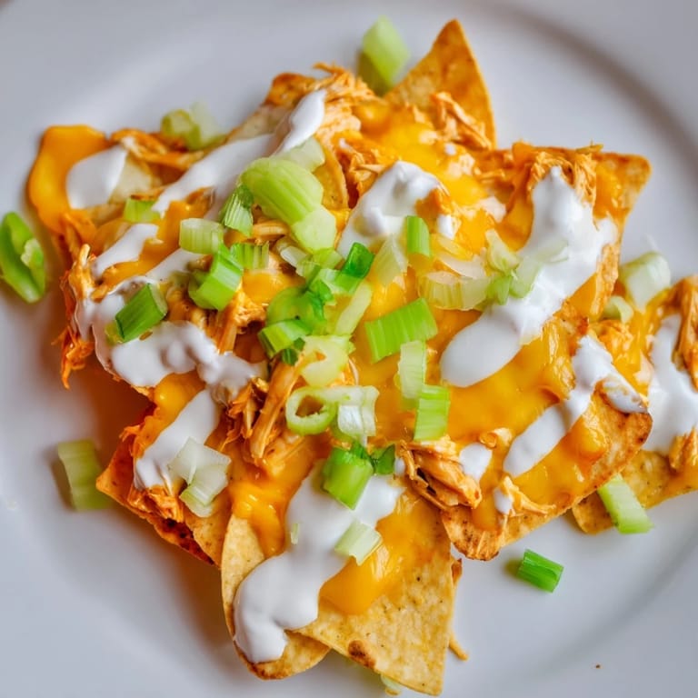 Freshly baked Buffalo chicken nachos topped with green onions, celery, and tomatoes, served steaming hot on a platter.  